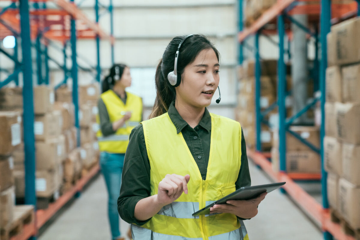 jeune femme d'affaires portant un casque d'écoute parlant avec l'entreprise et tenant une tablette numérique mobile regardant les marchandises en stock vérifiant le colis de commande. bokeh collègue féminine se tenant à l'arrière dans l'entrepôt.